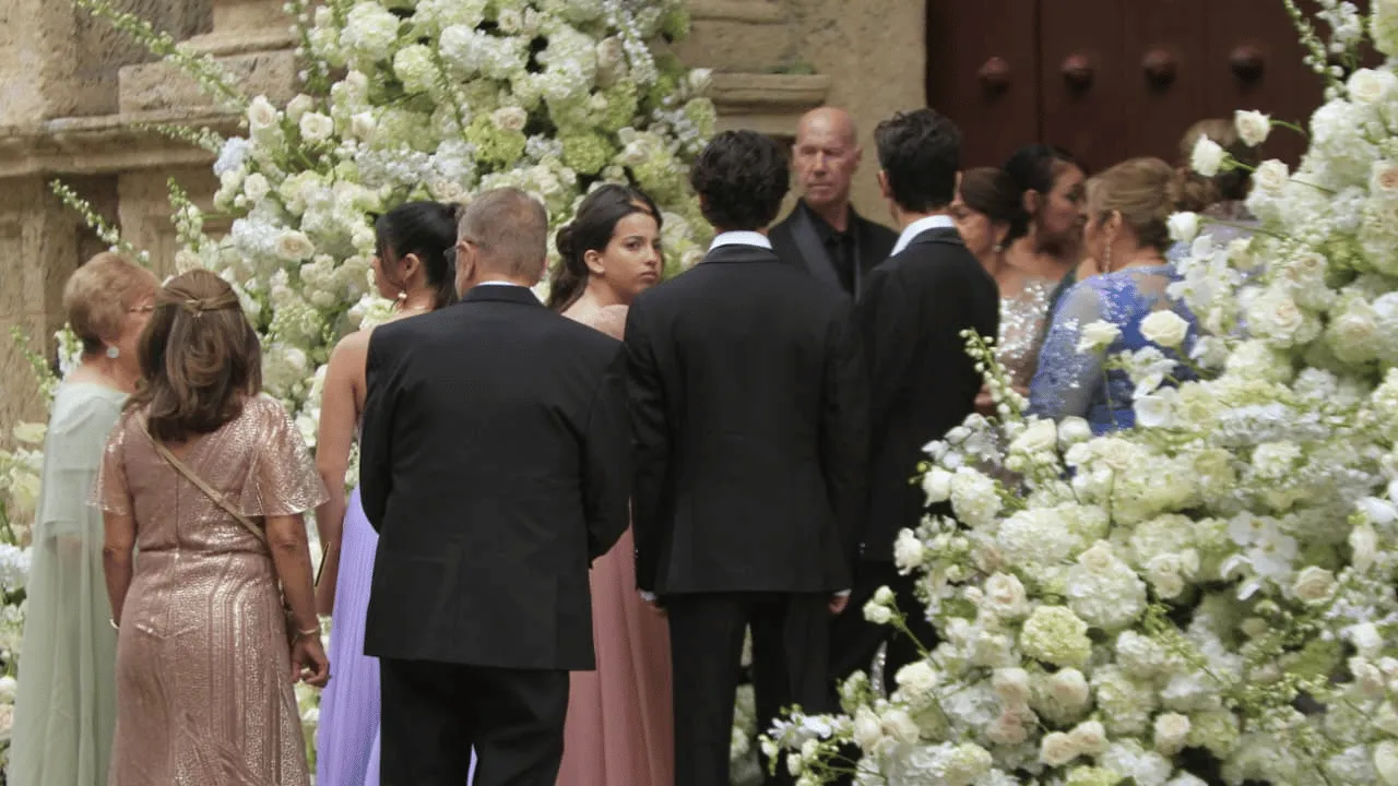 Invitados de traje y vestidos largos de colores esperan frente a arreglos de flores blancas.